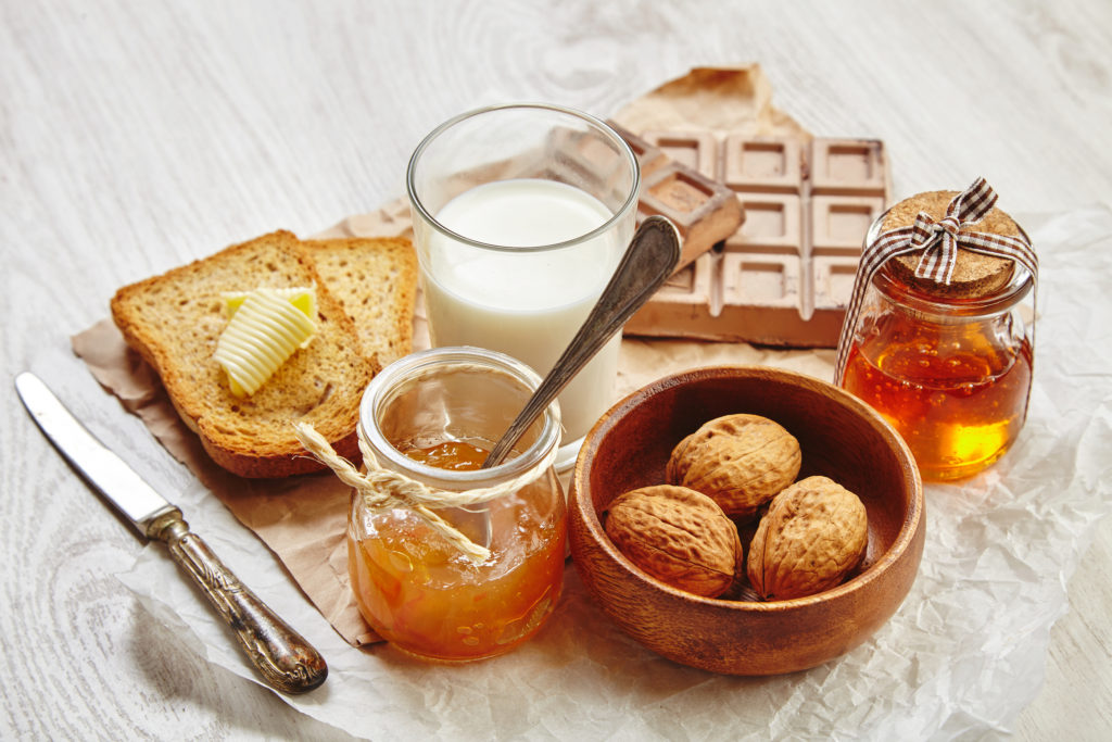 Side view of breakfast set with chocolate walnuts in wooden bowl jam honey in gift jar dry toast bread butter and milk. Everything on craft paper and vintage knife and spoon with patina.